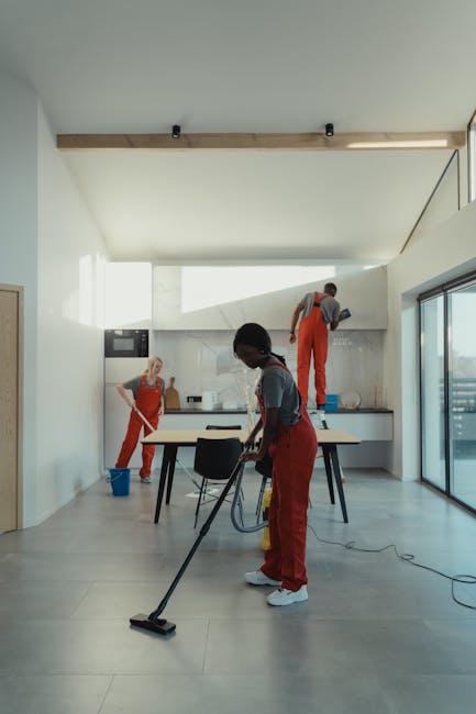 A professional cleaner with blonde hair, smiling and wearing a white blouse and black apron, engaged in surface cleaning during a domestic or commercial cleaning session at a well-lit window area. The