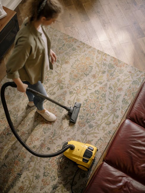 A woman using a yellow vacuum cleaner to deep clean a patterned, light-colored rug in a living room with wooden flooring and a burgundy leather sofa. The room is well-lit, and dust and dirt are being 