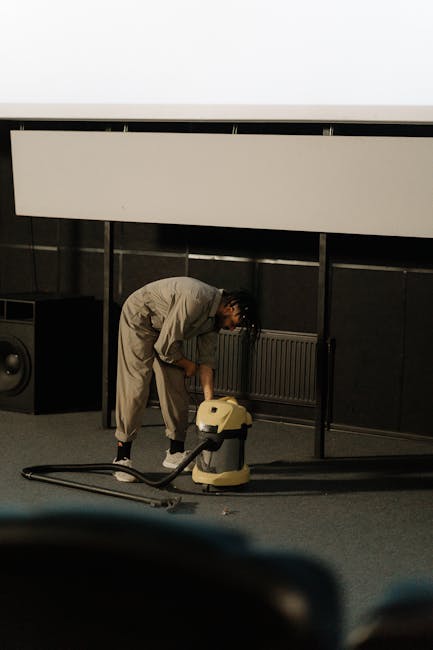 A woman using a yellow vacuum cleaner to deep clean a patterned, light-colored rug in a living room with wooden flooring and a burgundy leather sofa. The room is well-lit, and dust and dirt are being 