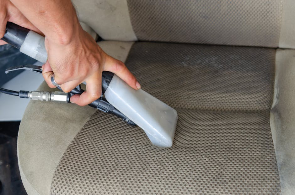 A woman with dark curly hair wearing a light grey T-shirt and red checkered pajama pants vacuuming a white carpet in a bright bedroom. The room features white walls, a large window with sheer white cu