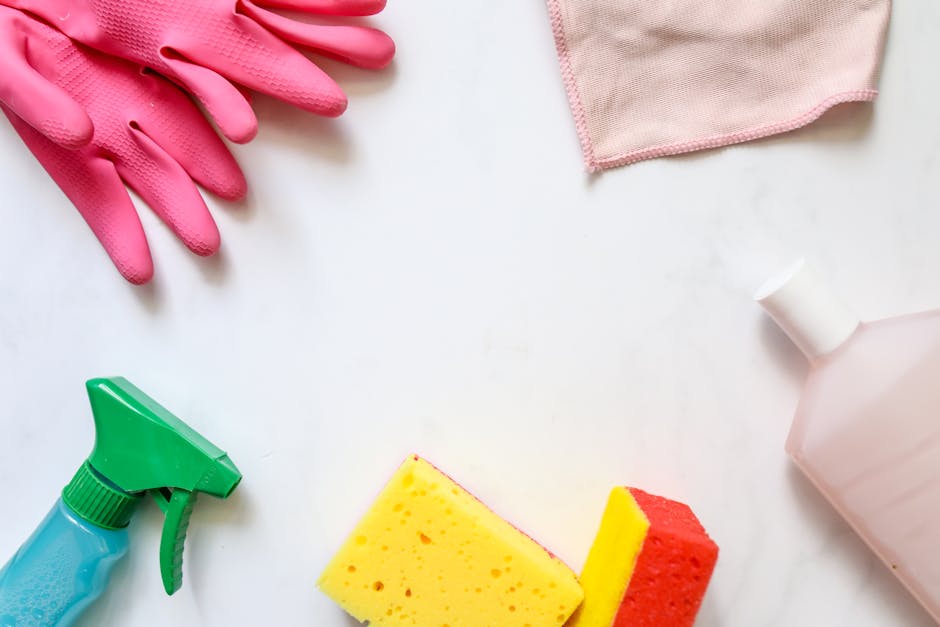 An arrangement of cleaning tools and supplies for surface cleaning and sanitisation, including a pair of pink rubber gloves positioned at the top left corner, a pink cloth in the top right corner, and